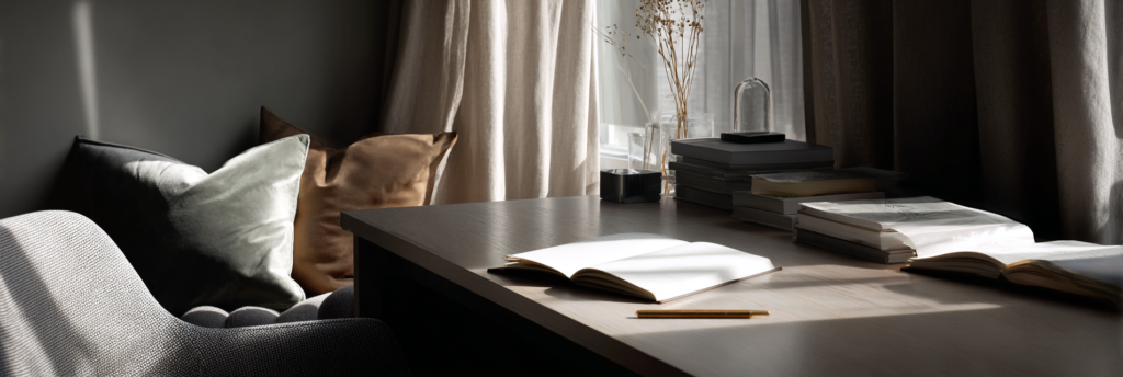 A quiet writing desk by a window with an open notebook, a pencil resting on the table, stacked books nearby, and soft daylight filtering through curtains.