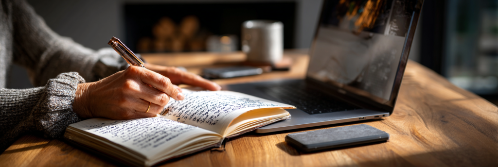 A close-up of a hand writing in a spiral notebook beside an open laptop, warm ambient light highlighting handwritten notes while the screen glows softly in the background.